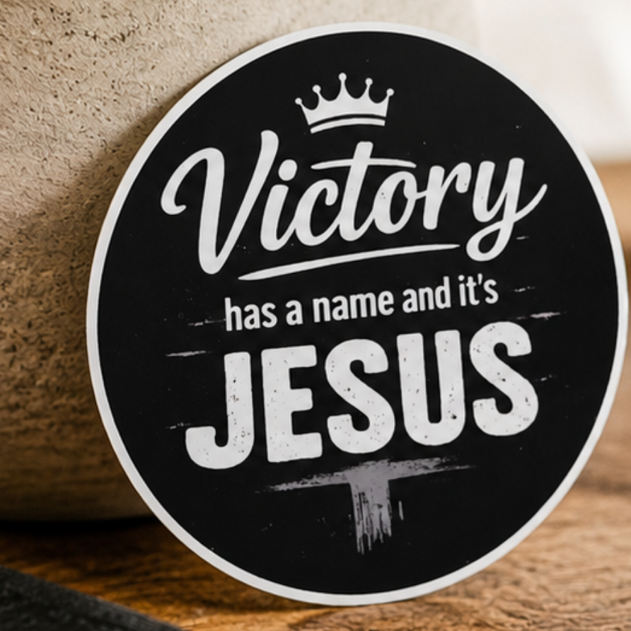 A round black sign with white text reading 'Victory has a name and it's JESUS' and a small crown at the top, displayed on a wooden surface next to a Bible and a potted plant.