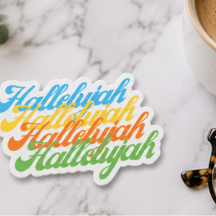 A die-cut sticker with the word 'Hallelujah' repeated in a colorful script, placed on a white marble surface next to a cup of coffee and tortoiseshell eyeglasses.